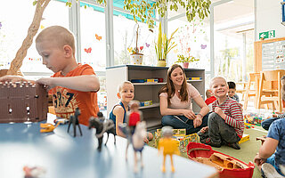 Ein Kind spielt mit Playmobil auf einem Tisch, im Hintergrund spielt eine Betreuerin mit einem Kind auf dem Boden