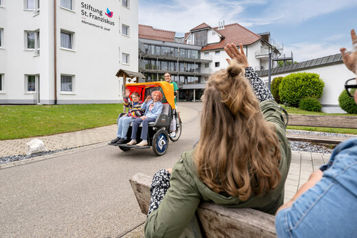 Zwei ältere Menschen sitzen in einer Fahrrad-Rikscha, die von einer Pflegeperson gefahren wird und winken zwei Menschen zu, die auf einer Bank sitzen