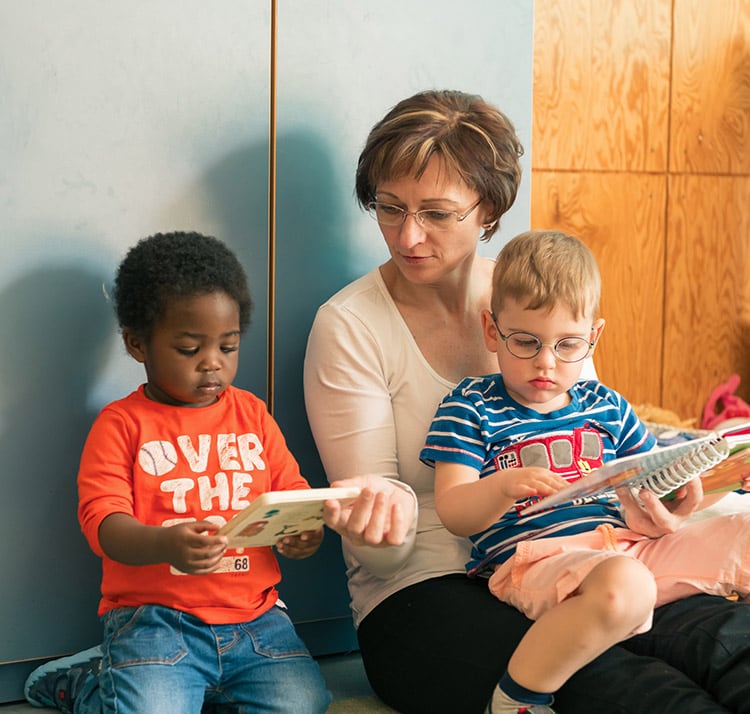 Kindergärtnerin sitzend unterhält sich mit zwei Kindern, die ein Bucj in der Hand halten, eines sitzt auf ihrem Schoß