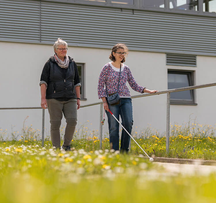 Mobilitätstrainerin mit junger Frau mit Sehbehinderung, die mit einem Langstock im Gelände läuft