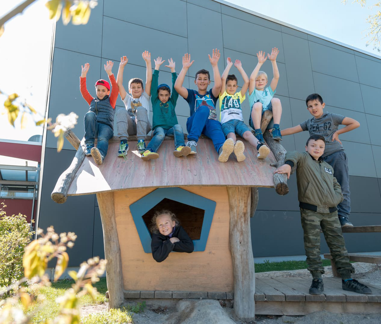Kinder auf einem Holz-Spielhaus sitzend reißen die Hände in den Himmel 