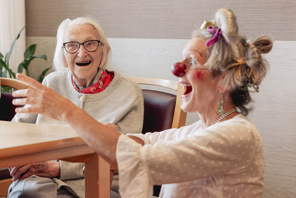 Ältere Dame am Tisch sitzend lacht mit einer Frau, die einen Clown spielt